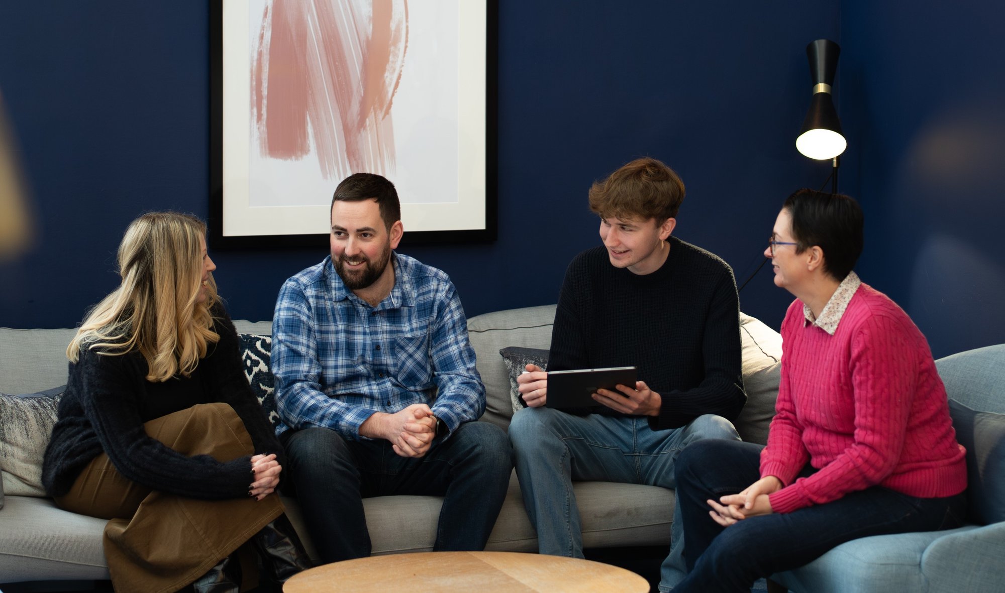 Two men and two women are sat on a sofa talking in a business setting.