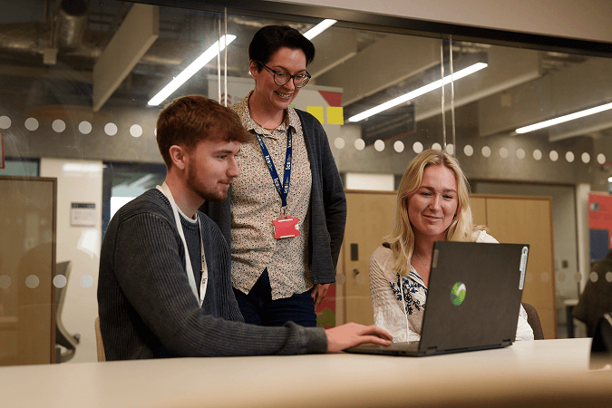A man and a woman looking at a laptop. A smiling woman overlooks them while standing.